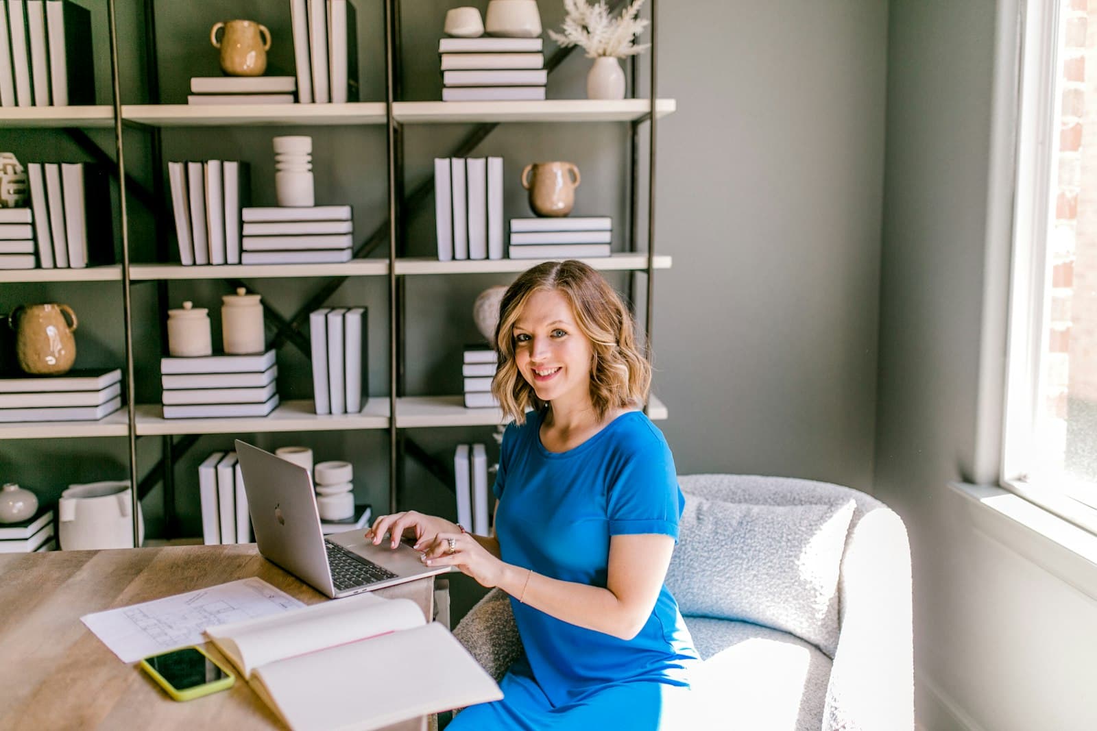 A smiling woman working on a laptop at a desk, with a notebook and printed plans beside her