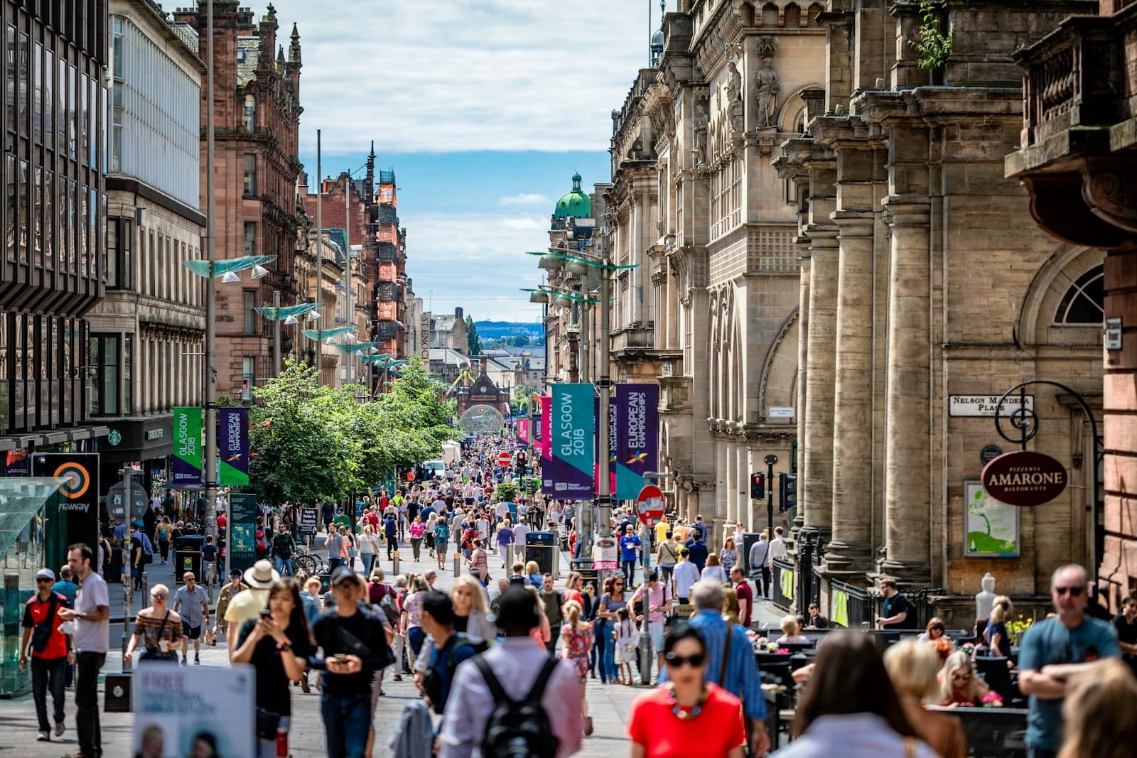 Shoppers walking down Buchanan Street in Glasgow on a sunny day, between red sandstone buildings, with street banners and trees along the pedestrian street