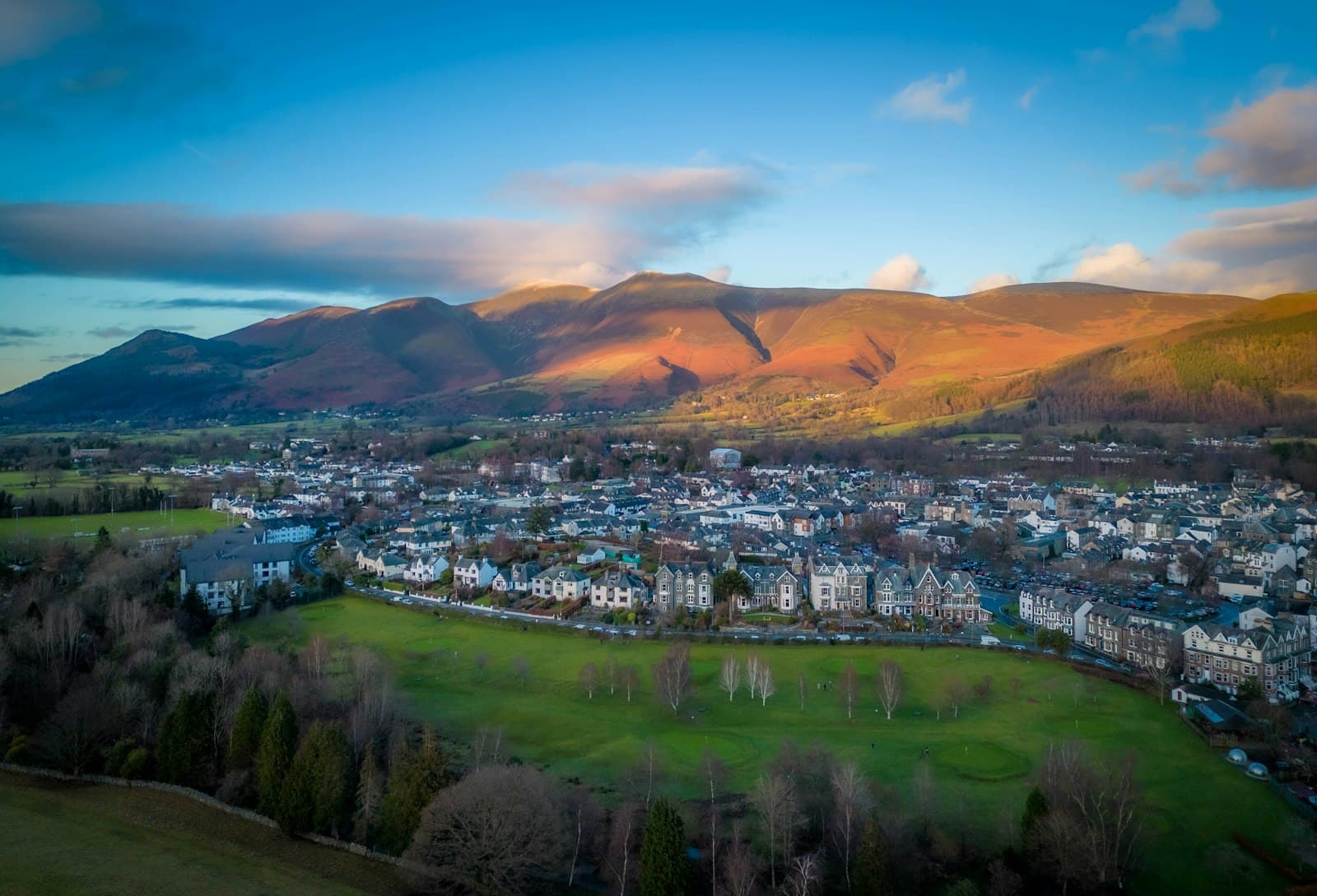 Aerial view of Keswick in Cumbria at sunset, rows of grey slate houses beside a green park, with the fells behind lit orange under a blue sky