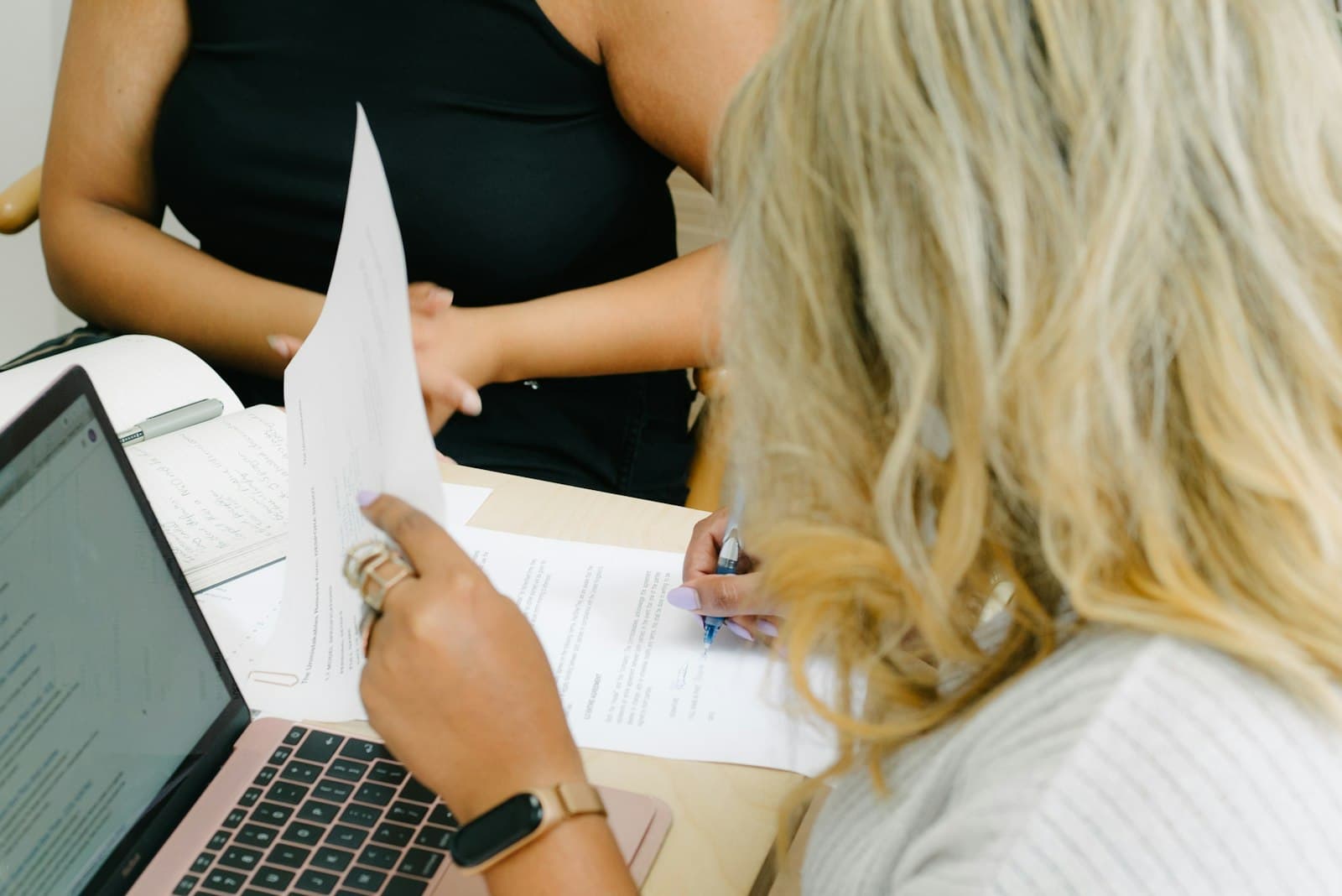 Two women at a desk going through printed papers beside an open laptop