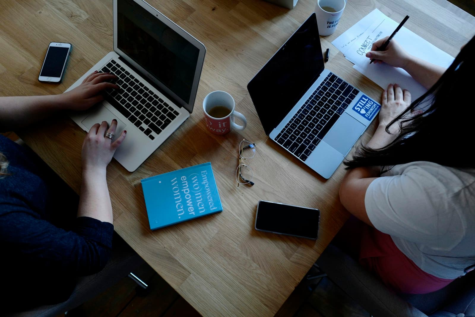 Overhead view of two people working on laptops at a wooden table with mugs and a notebook