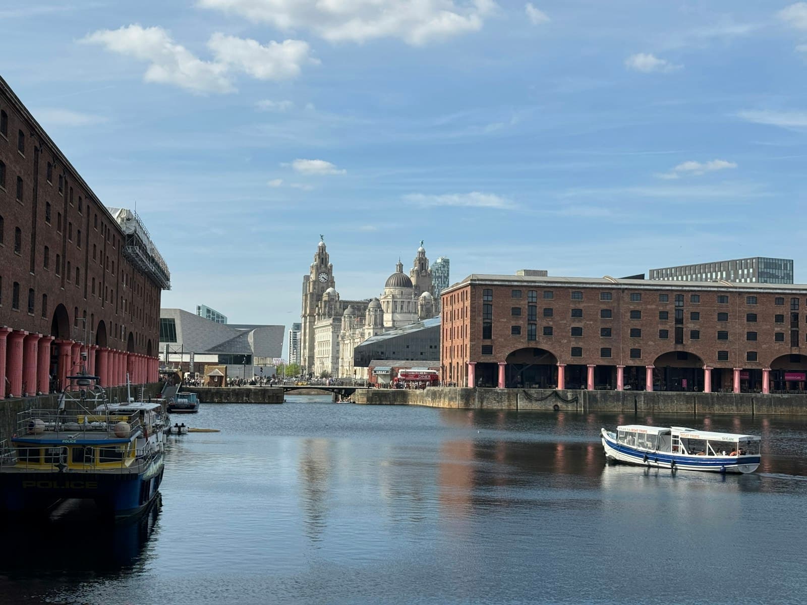 Royal Albert Dock in Liverpool on a sunny day, red-brick warehouses on both sides of the water, a small ferry boat on the dock and the Royal Liver Building in the distance under a blue sky