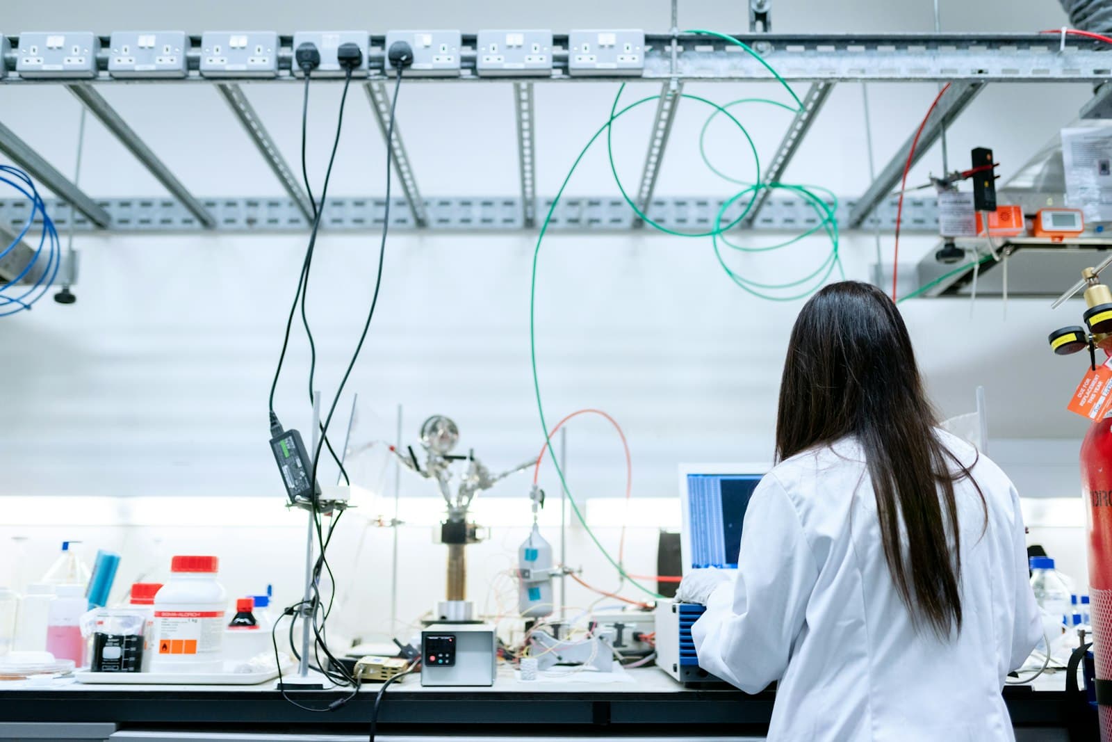 A scientist in a white lab coat, seen from behind, working at a laboratory bench of glassware, reagent bottles and wired test equipment, under an overhead rail of UK three-pin sockets