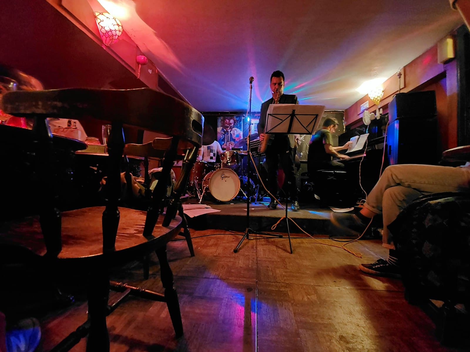 A jazz trio playing on the small low stage of a London jazz club, a saxophonist at the microphone behind a music stand, a drummer and a pianist beside him, seen from among the wooden pub chairs under red and blue lights