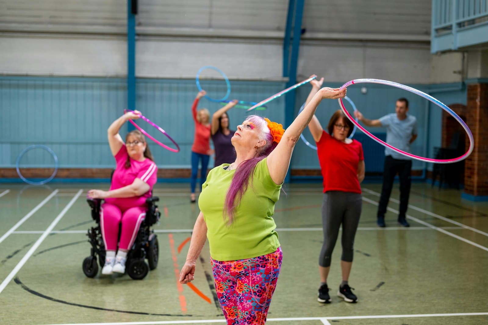 A mixed-ability hula hooping session in a sports hall, including a wheelchair user, representing UK disability organisations