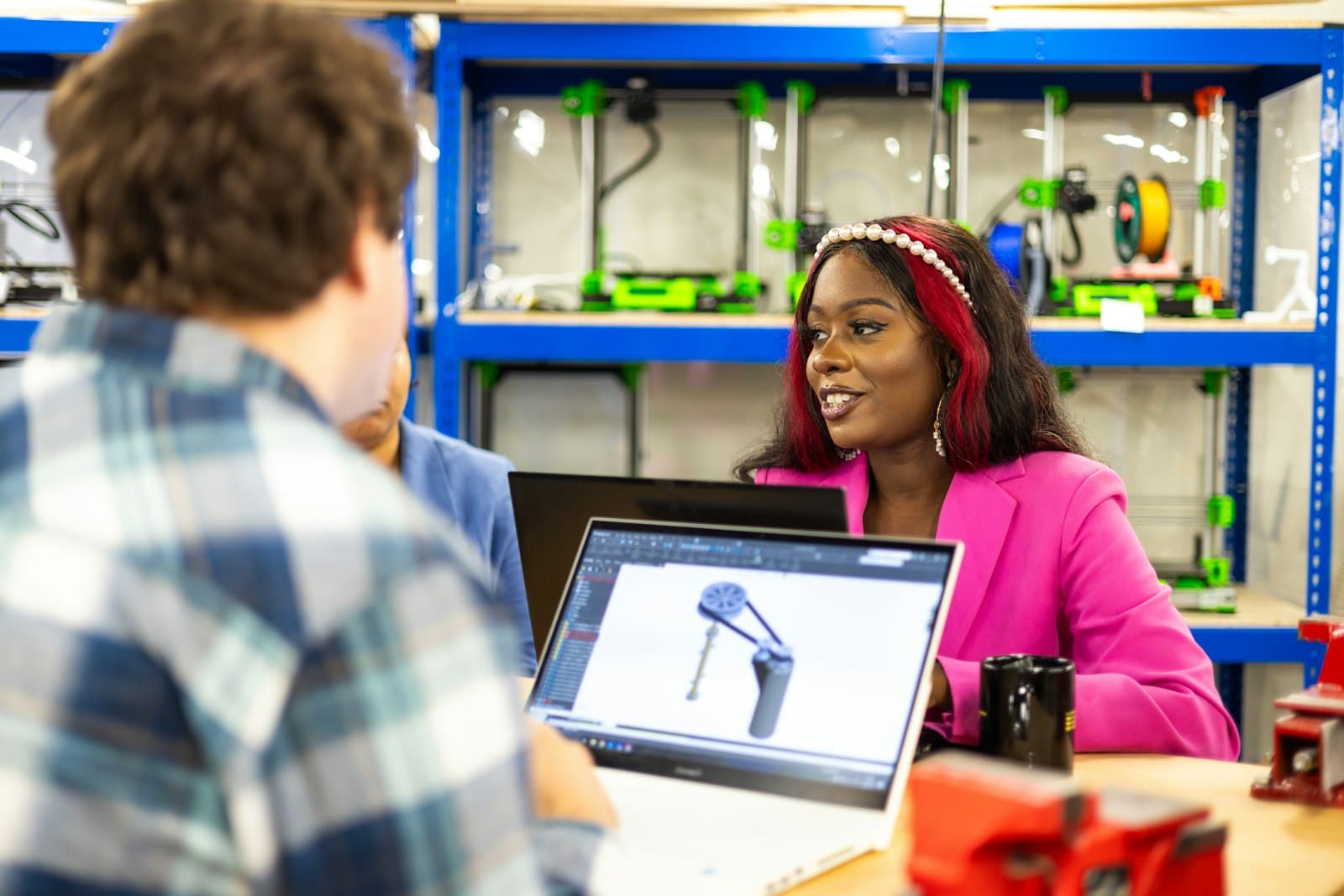 A woman in a pink blazer discussing a 3D product design on a laptop across a workshop table, with 3D printers on the shelves behind