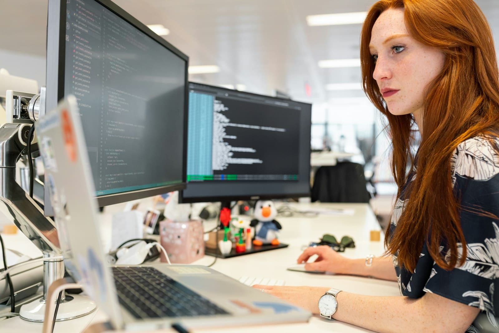 A software engineer with long red hair working at her desk, lines of code on two monitors and an open laptop in front of her