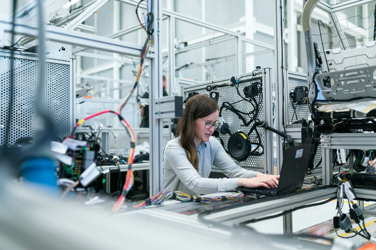 An engineer in glasses working on a laptop in a vehicle test lab, surrounded by wiring looms, a car door panel and a seat frame on metal test rigs