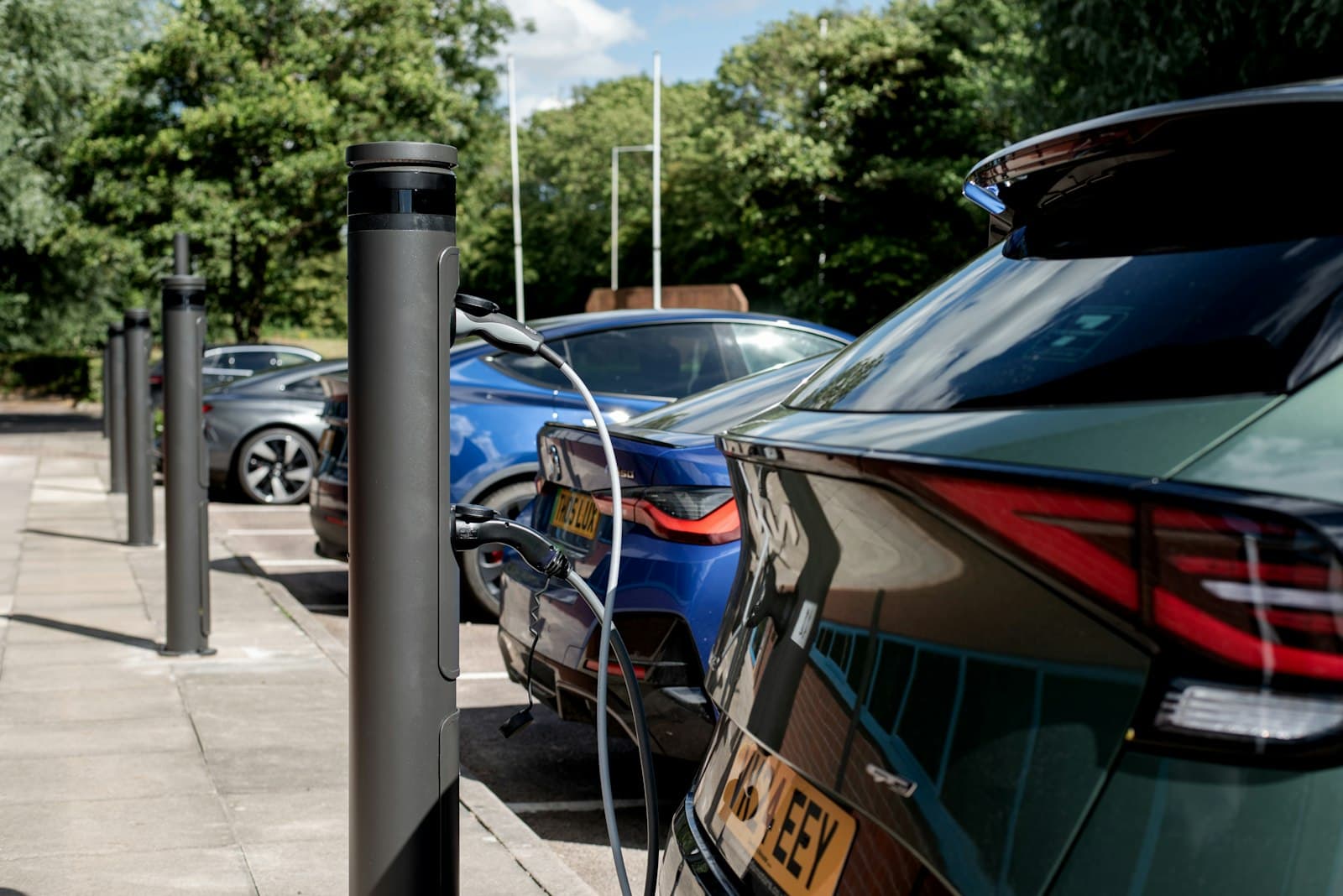 A row of electric cars plugged into tall black charging posts in a sunny workplace car park, with UK yellow number plates and trees behind