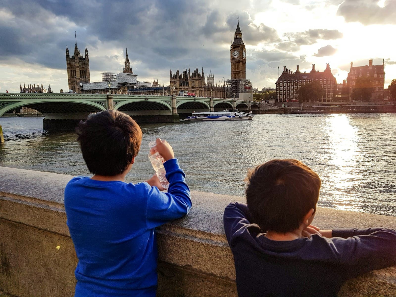 Two boys in blue tops leaning on the Thames embankment wall, looking across the river at Westminster Bridge, the Houses of Parliament and Big Ben, a river boat passing under a cloudy late-afternoon sky
